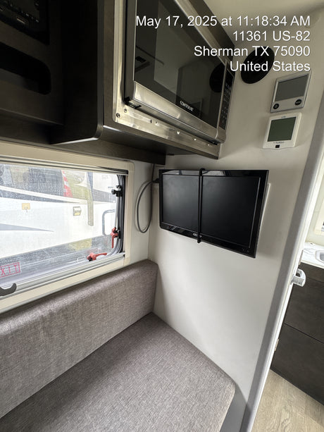 View of a modern rv rental interior: gray upholstered seating, wall-mounted TV, microwave, and window showing exterior.