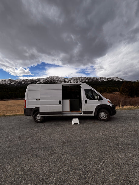 White RV rental camper van with open side door, step stool, parked by snowy mountains under a cloudy sky.