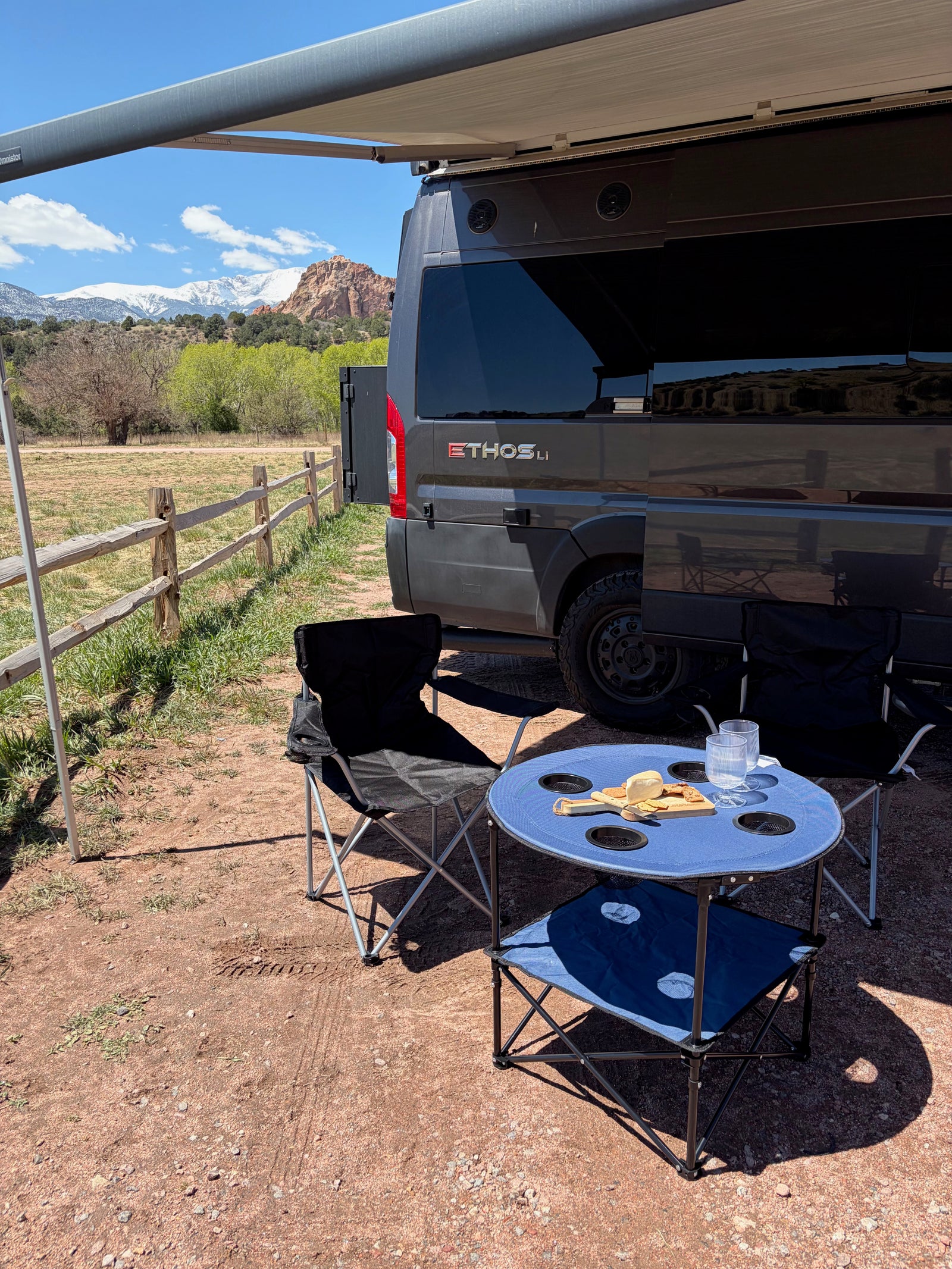 An RV rental camper van with an awning out, a blue picnic table, chairs, and snacks, facing snow-capped mountains.