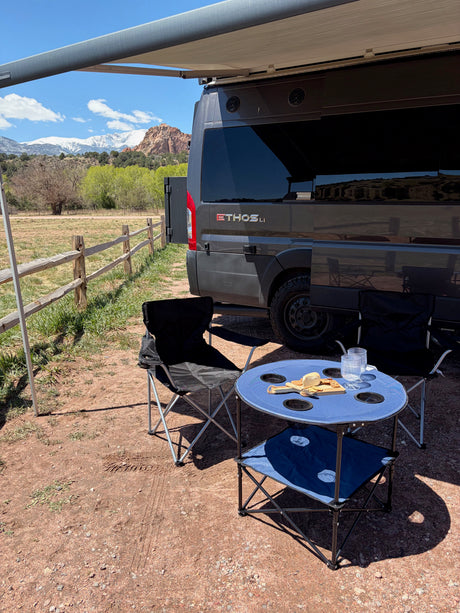 An RV rental camper van with an awning out, a blue picnic table, chairs, and snacks, facing snow-capped mountains.