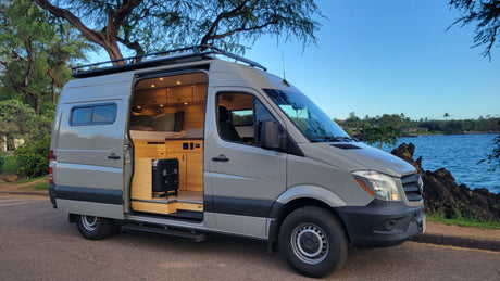 Gray rv rental van with an open door, revealing a warm, wooden interior. Parked by a rocky ocean shore under blue sky.