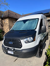 A custom white Ford Transit van with a black hood & rugged tires, parked. Solar panels are visible on the house behind it. Great for an RV rental.