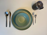 RV rental dining setup: teal-green plates, complete cutlery (spoon, knife, fork), and a handy travel mug on a white surface.