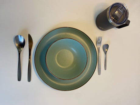 RV rental dining setup: teal-green plates, complete cutlery (spoon, knife, fork), and a handy travel mug on a white surface.