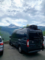 A dark grey Thor Tellaro RV rental is parked at a mountain overlook, with a majestic snow-capped peak and cloudy sky.
