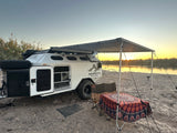 Mammoth Overland RV rental set up riverside at sunset, featuring a travel trailer with awning, table, and chair.