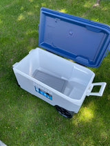 Open white Igloo wheeled cooler on green grass, blue lid beside it, showing gray tray inside. Ideal for your RV rental.