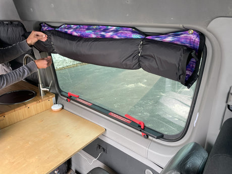 RV rental interior: Person lowers an insulated window curtain, showcasing the kitchenette, and emergency exit handles.