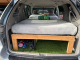 RV rental ready: van interior with a custom bed platform, mattress, pillows, and artificial turf storage underneath.