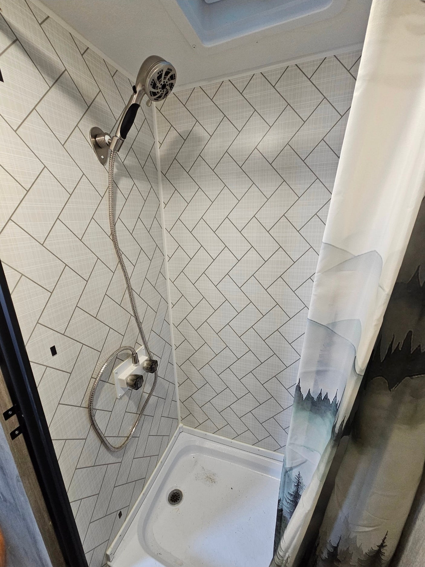 Compact RV rental shower interior: herringbone walls, flexible shower head, and a nature-themed curtain.