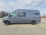 This gray Sprinter camper van, featuring a roof rack, is ideal for an RV rental, parked in a lot under cloudy skies.
