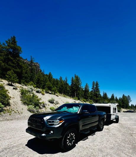 Toyota Tacoma truck towing a compact RV rental trailer on a dirt road, amidst rocky hills & pine trees, clear blue sky.