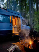 Cozy RV rental van in a dark forest at night, warmly lit inside with open door, beside a glowing campfire and chairs.