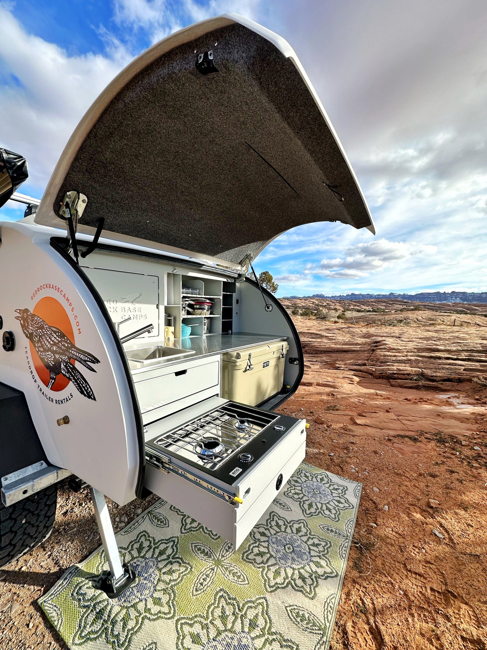 Teardrop camper RV rental outdoor kitchen with sink, stove, and cooler, set up in a red rock desert.