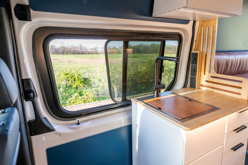 View from inside an RV rental showing a modern kitchen area with a sink, wood counter, and a serene green field outside the window. — photo 6