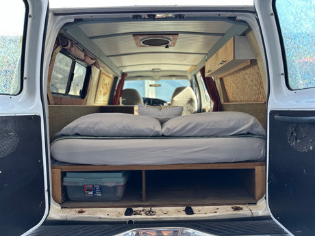 RV rental: Inside a cozy camper van conversion with a bed, under-bed storage, overhead cabinets, and a ceiling fan.
