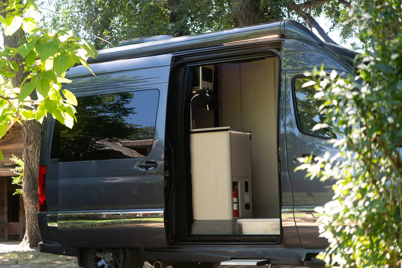 An open door reveals the modern interior of this dark gray RV rental, featuring light wood cabinetry, a sink, and a fire extinguisher. — photo 8