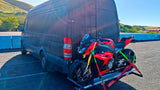 This customized dark grey Mercedes Sprinter van, an RV rental option, transports a red BMW S1000RR motorcycle on its rear carrier.
