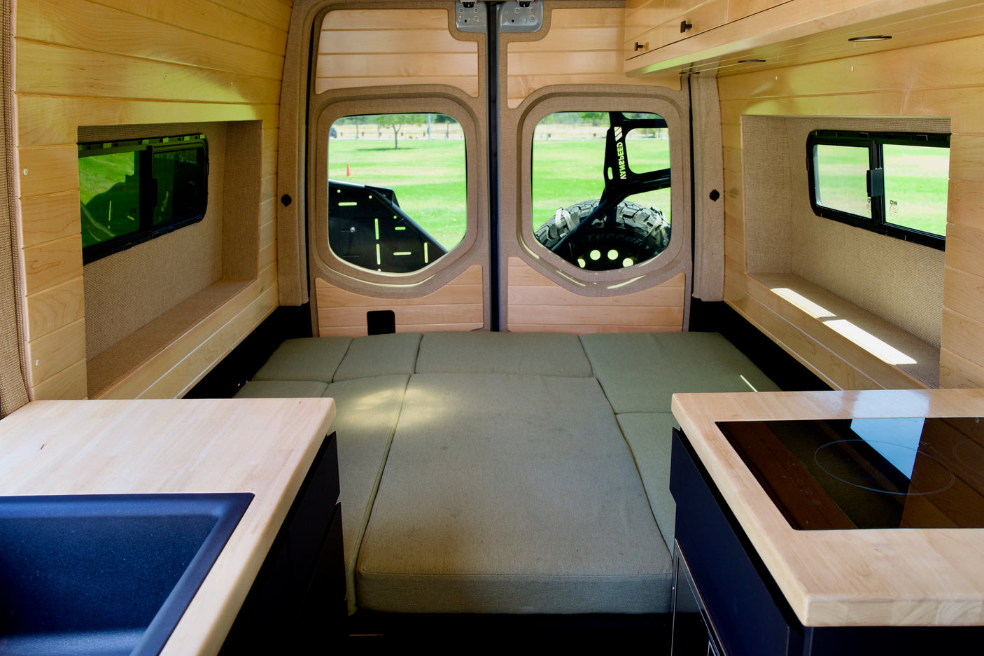 Cozy RV rental interior: a bed, kitchen with sink & stovetop, wood paneling, and rear windows revealing a green field.