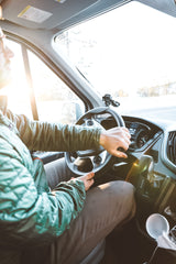 Inside an RV rental, a person in a green jacket drives, hands on the wheel. Bright sun illuminates the interior winter scene.