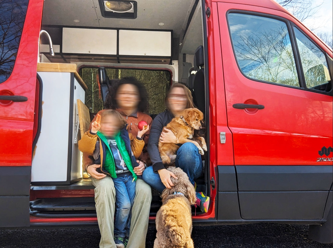 Happy family with child & two dogs pose by their red Sprinter RV rental, showcasing cozy van life travel.