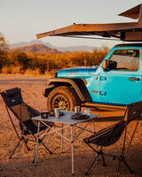 Perfect RV rental camping setup! Blue Jeep with rooftop tent, portable table, and chairs in a scenic desert.