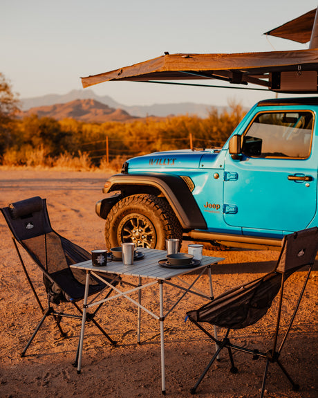 Perfect RV rental camping setup! Blue Jeep with rooftop tent, portable table, and chairs in a scenic desert.
