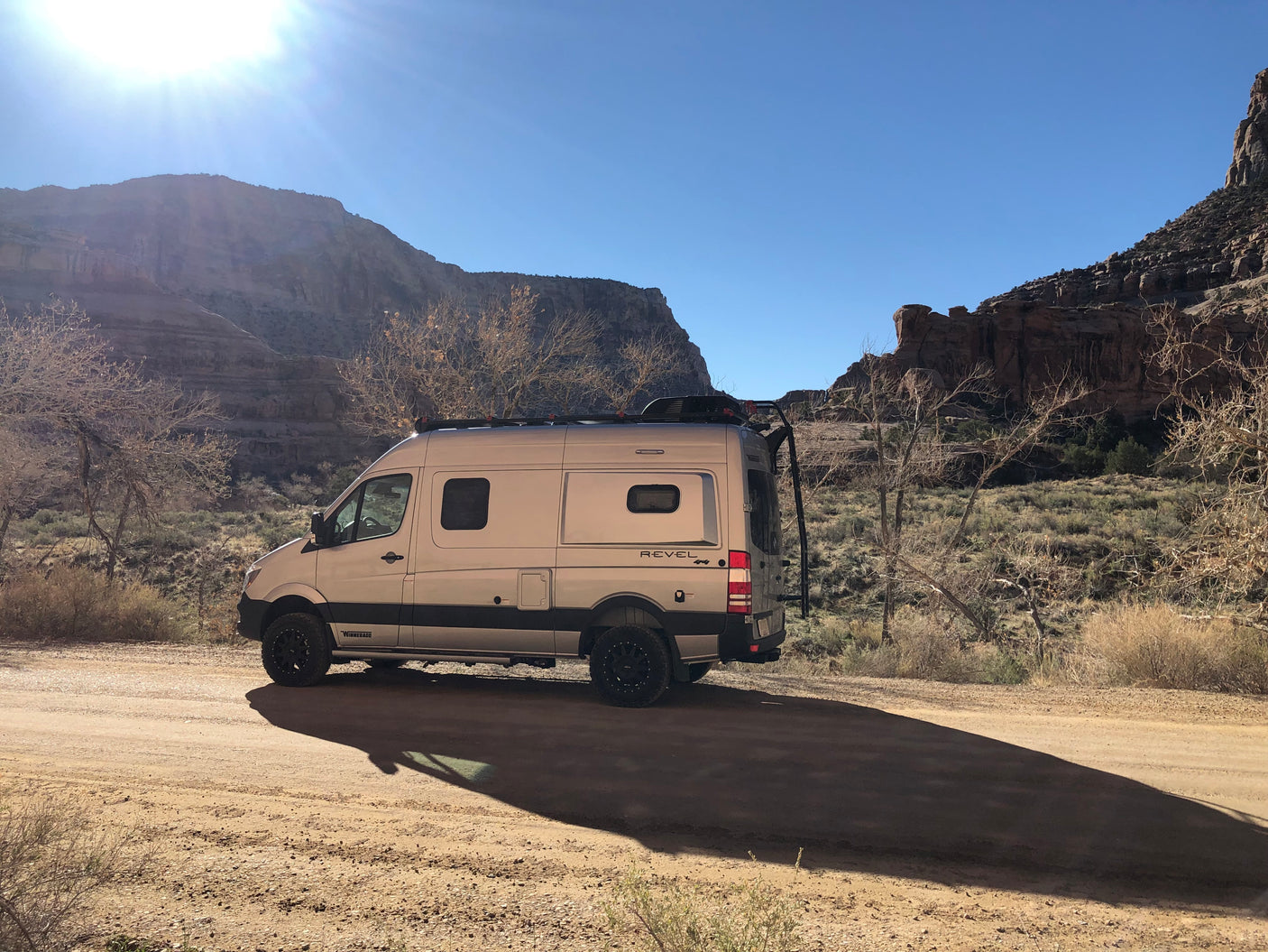 Winnebago Revel 4x4 rv rental parked on a desert dirt road with mountains under a sunny sky, casting a long shadow.