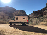Winnebago Revel 4x4 rv rental parked on a desert dirt road with mountains under a sunny sky, casting a long shadow.