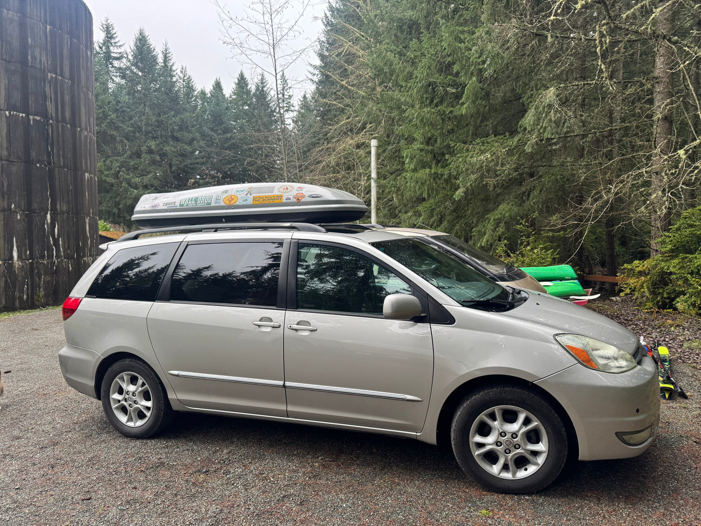 A silver minivan with a sticker-laden roof box in a woodsy setting, ideal for a memorable RV rental adventure.