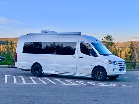 White Mercedes Sprinter RV rental camper van parked in a lot with a forested mountain backdrop. Ready for your next travel adventure!