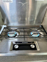 An RV rental two-burner gas stove with lit blue flames, a Suburban brand, control knobs, and a visible safety warning.