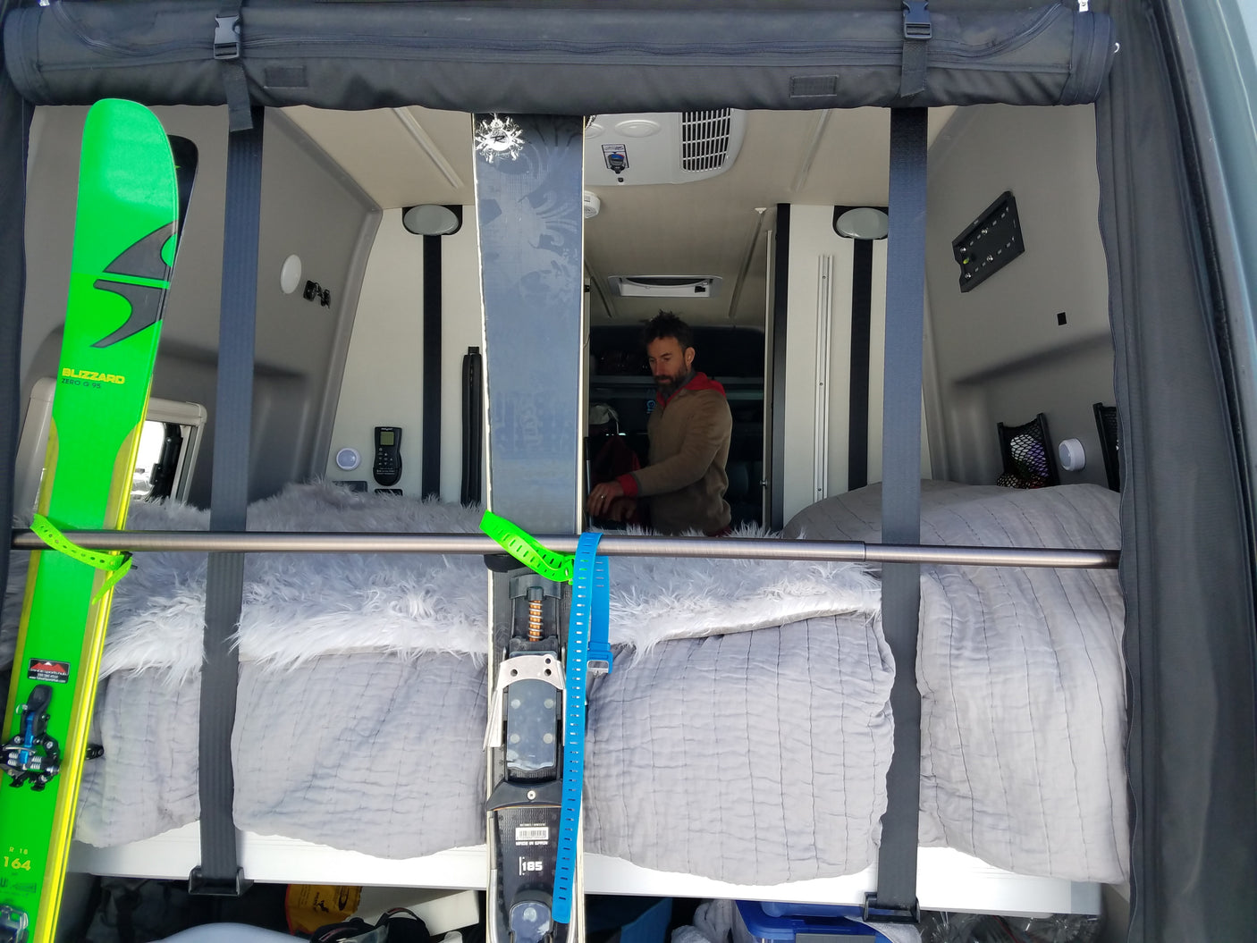 Inside an **rv rental**, a man sits in a camper van with neatly stored skis. Features comfortable beds and practical storage for winter sports gear.