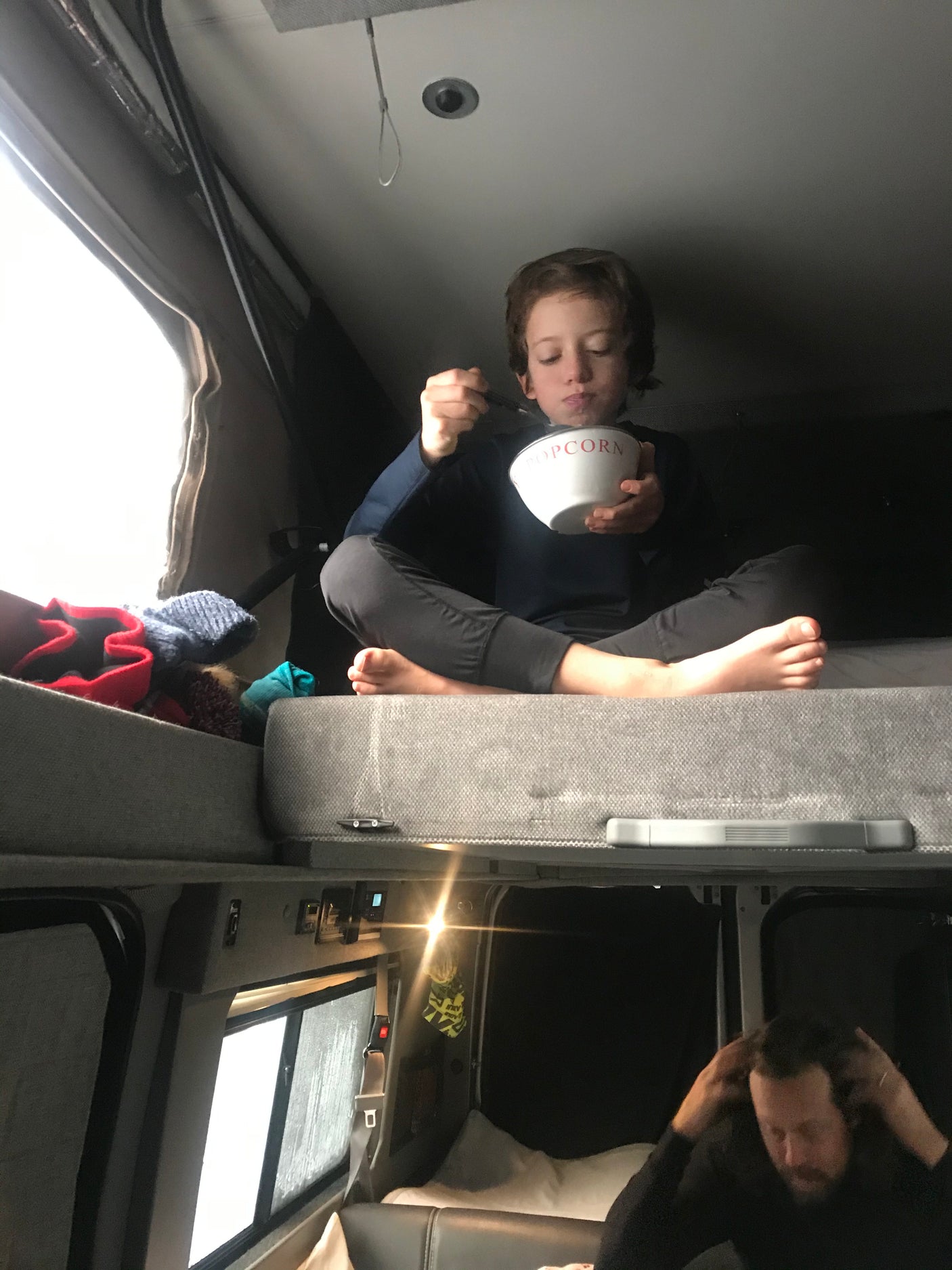 Inside a cozy RV rental: Child on upper bunk eating popcorn, man relaxing below. Ideal for family road trips.