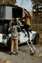 Setting up their RV rental roof top tent on a Jeep. A man helps a woman on a ladder, ready for outdoor adventure.