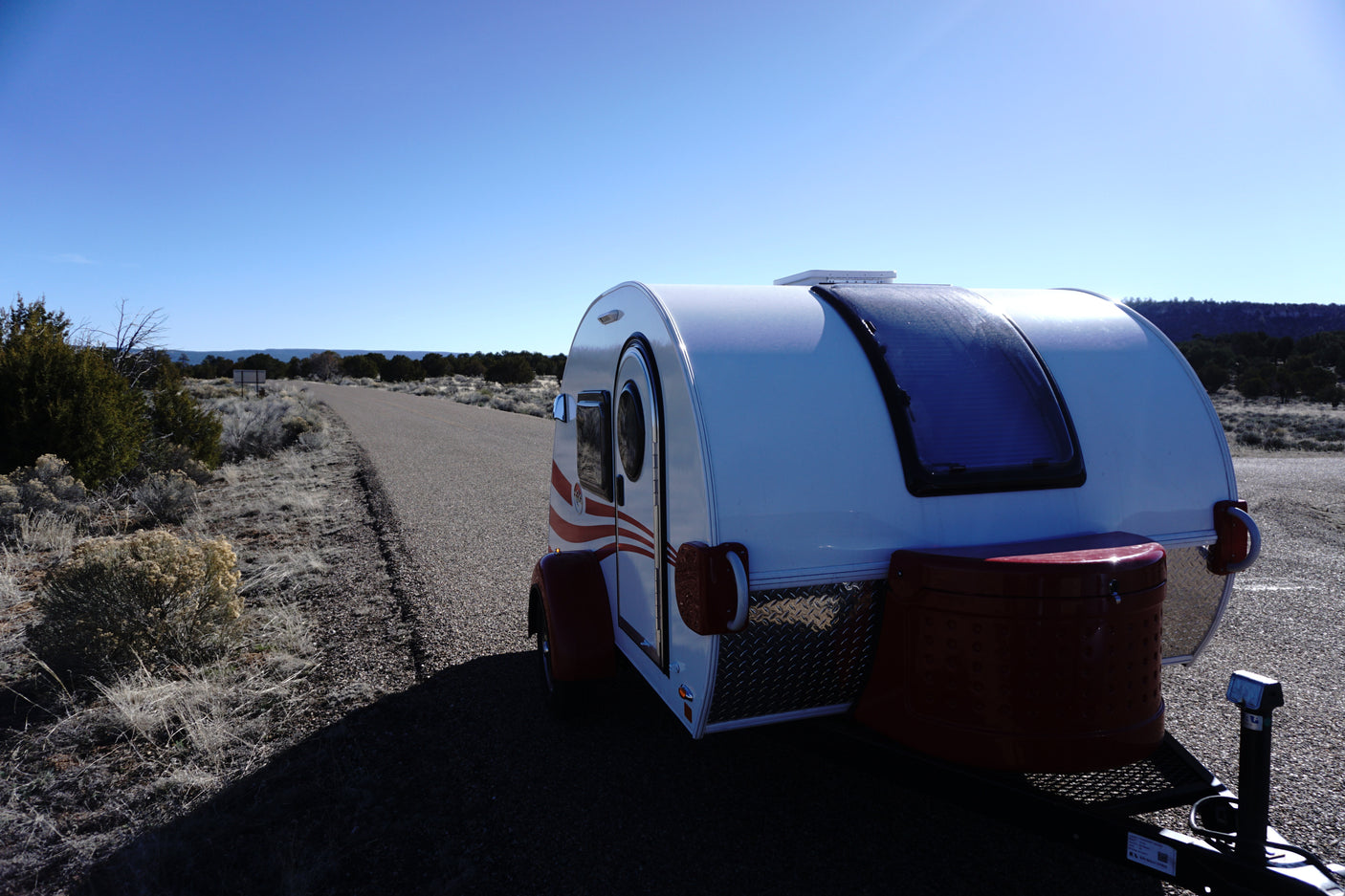 This white teardrop camper RV rental is ready for adventure on a desert road under a clear blue sky.