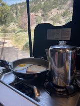 Breakfast cooking in an RV rental: egg, toast, and coffee on a camping stove, with a scenic outdoor view.