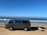 Dark gray RV rental van parked beachside with open hatch & surfboard. Surfers dot the ocean under a clear blue sky.