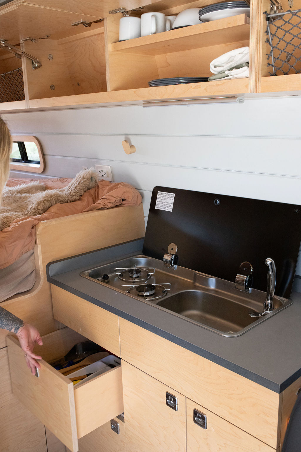 RV rental kitchen: compact stove/sink, open utensil drawer, overhead cabinets with dishes, and a cozy bed visible.