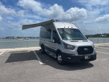 Silver Ford Transit camper van RV rental with extended awning, parked at a waterfront.