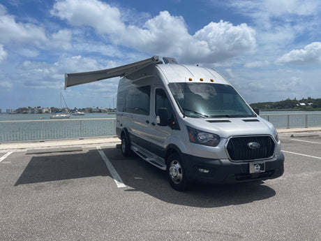 Silver Ford Transit camper van RV rental with extended awning, parked at a waterfront.