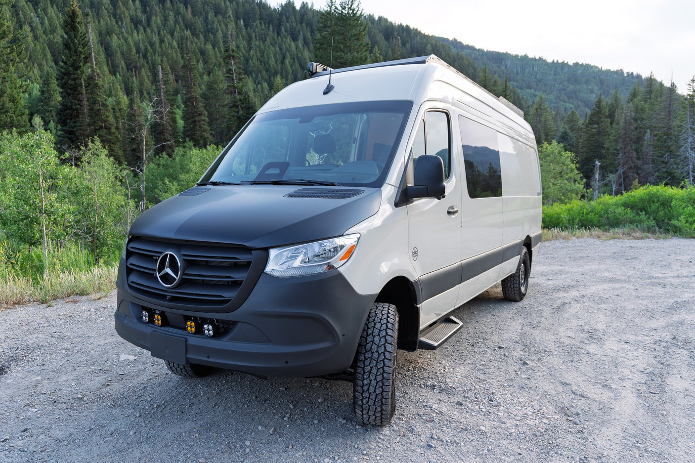 Rugged white Mercedes-Benz Sprinter camper van, ideal for rv rental, parked on gravel against a lush forest backdrop.