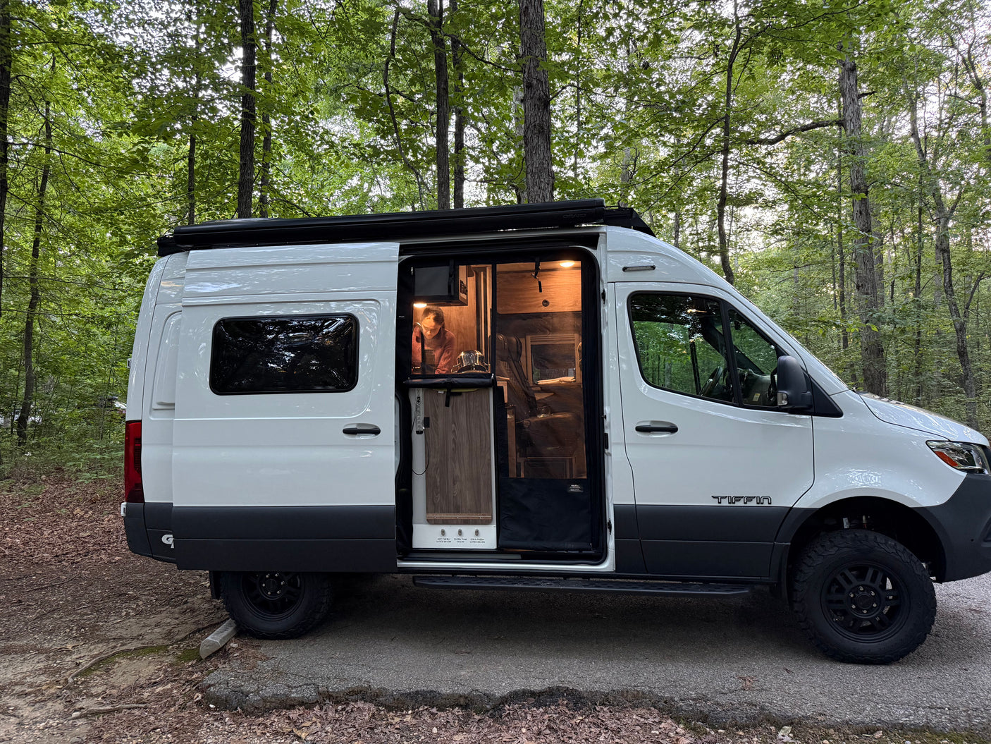Enjoying an RV rental adventure. A Tiffin camper van with its door open, showing a woman inside working, parked in a forest.