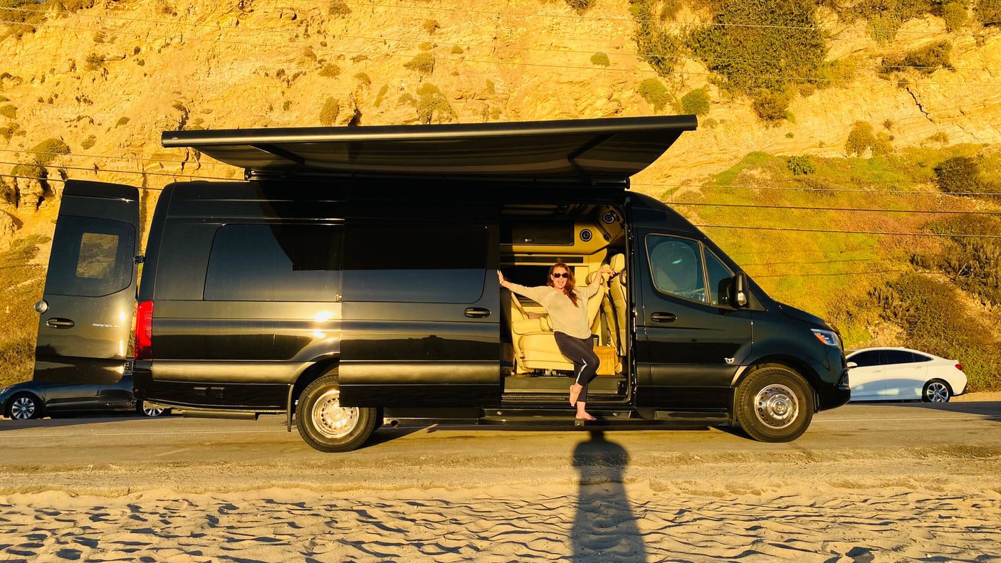 Barefoot woman smiles from her black luxury RV rental van, parked by a sunny beach and cliffs.