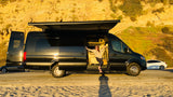 Barefoot woman smiles from her black luxury RV rental van, parked by a sunny beach and cliffs.