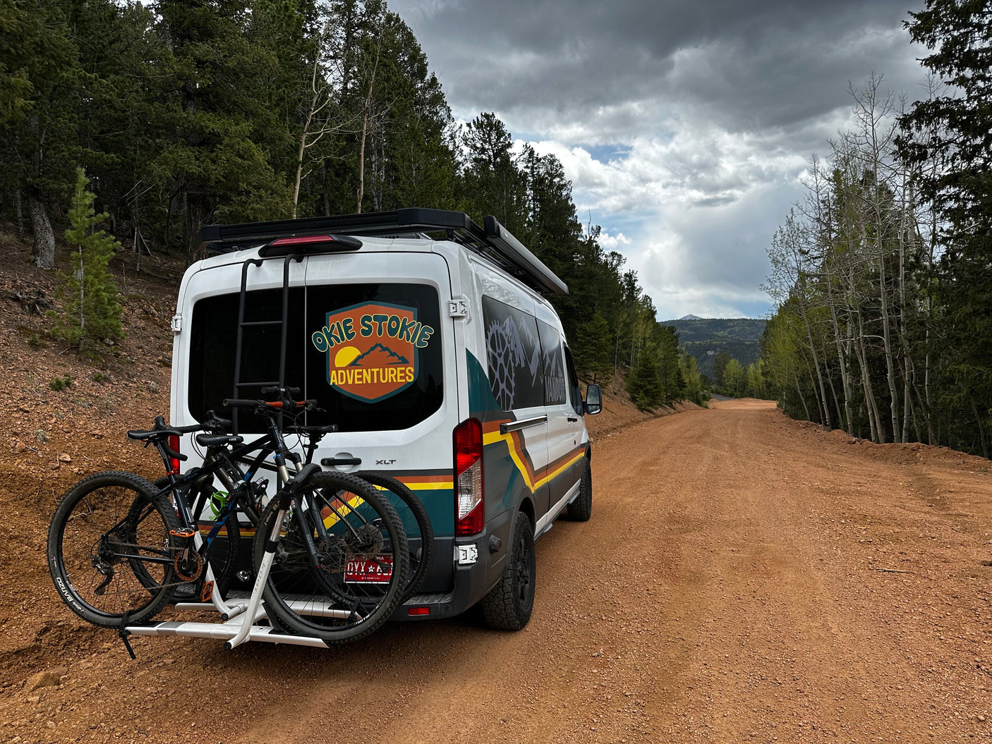 "Okie Stokie Adventures" RV rental van with mountain bikes parked on a gravel forest road.