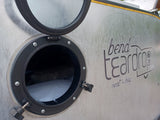 Side view of a Bend Teardrop RV rental with its round window open, revealing a cozy bed inside.