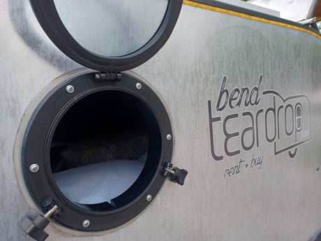 Side view of a Bend Teardrop RV rental with its round window open, revealing a cozy bed inside.