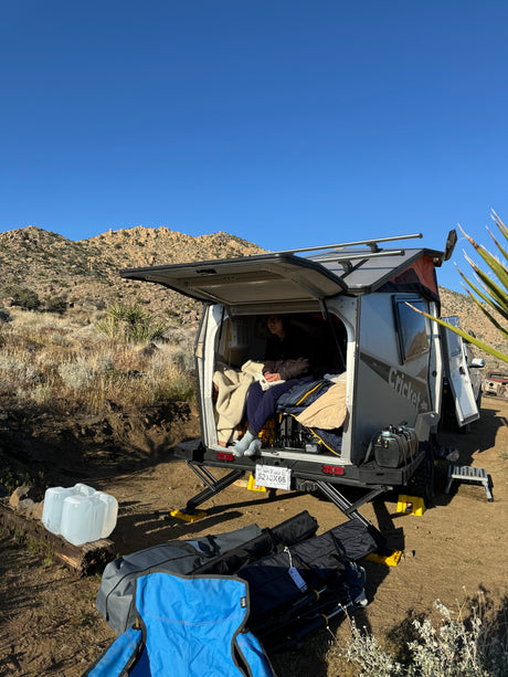 A person relaxes inside a small 'Cricket' camper, a perfect RV rental for a desert mountain camping adventure.