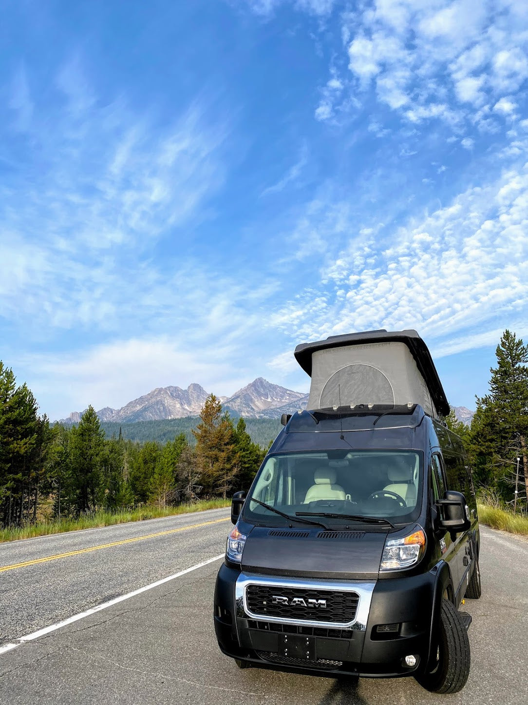 Experience nature with an RV rental! Dark camper van, pop-top up, on a scenic road with mountains & blue sky.